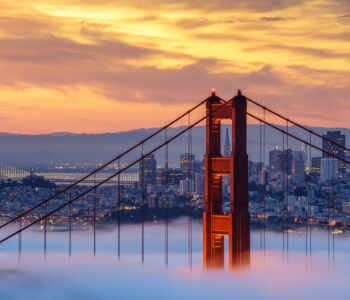 Golden Gate Bridge tower emerging from low coastal fog at sunset, with downtown San Francisco in the background under a glowing orange sky.