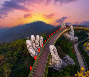 A panoramic aerial view of Da Nang’s Golden Bridge at sunrise, with massive stone hands lifting a curved wooden walkway above lush green mountains.