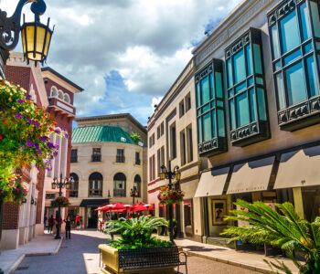 Rodeo Drive shopping street with upscale boutiques, flower-lined lampposts, and European-style architecture under a bright sky.