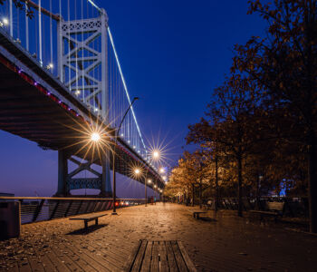 Night view from Race Street Pier with the Ben Franklin Bridge illuminated above a quiet waterfront walkway.
