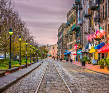 Cobblestone street lined with historic brick buildings and riverside shops along Savannah’s River Street at dusk.