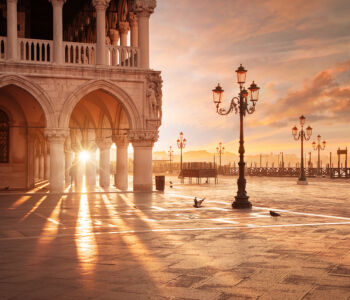 Golden sunrise casting long shadows across St. Mark’s Square, with ornate columns and lampposts glowing in the early light of Venice.