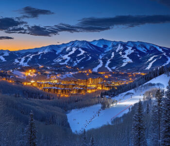 Aspen Snowmass Ski Resort at dusk with glowing lights and snow-covered slopes surrounded by mountain peaks in Colorado.