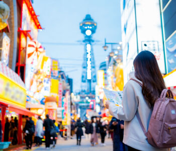 Traveler exploring Shinsekai district in Osaka, Japan, with Tsutenkaku Tower glowing in the background at dusk