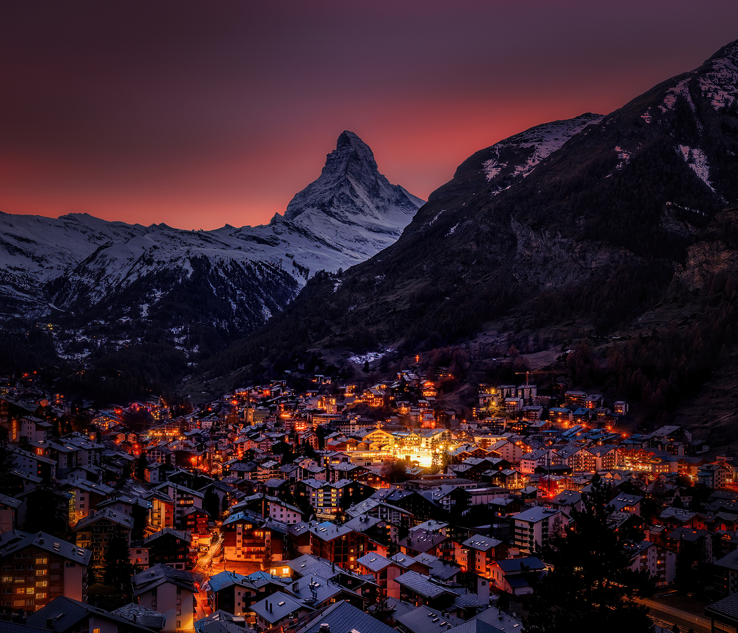 Twilight view of Zermatt, Switzerland with glowing chalet lights beneath snow-covered peaks and the Matterhorn in the distance