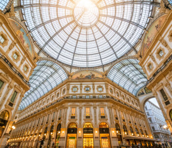 Interior view of the Galleria Vittorio Emanuele II in Milan, showcasing its grand glass-and-iron dome, ornate façades, and golden sunlight streaming through the historic arcade.
